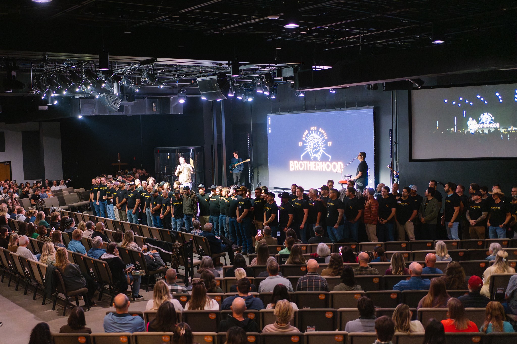 Men in black Brotherhood shirts lined across a stage before a congregation, a glowing Born for Battle screen behind them.