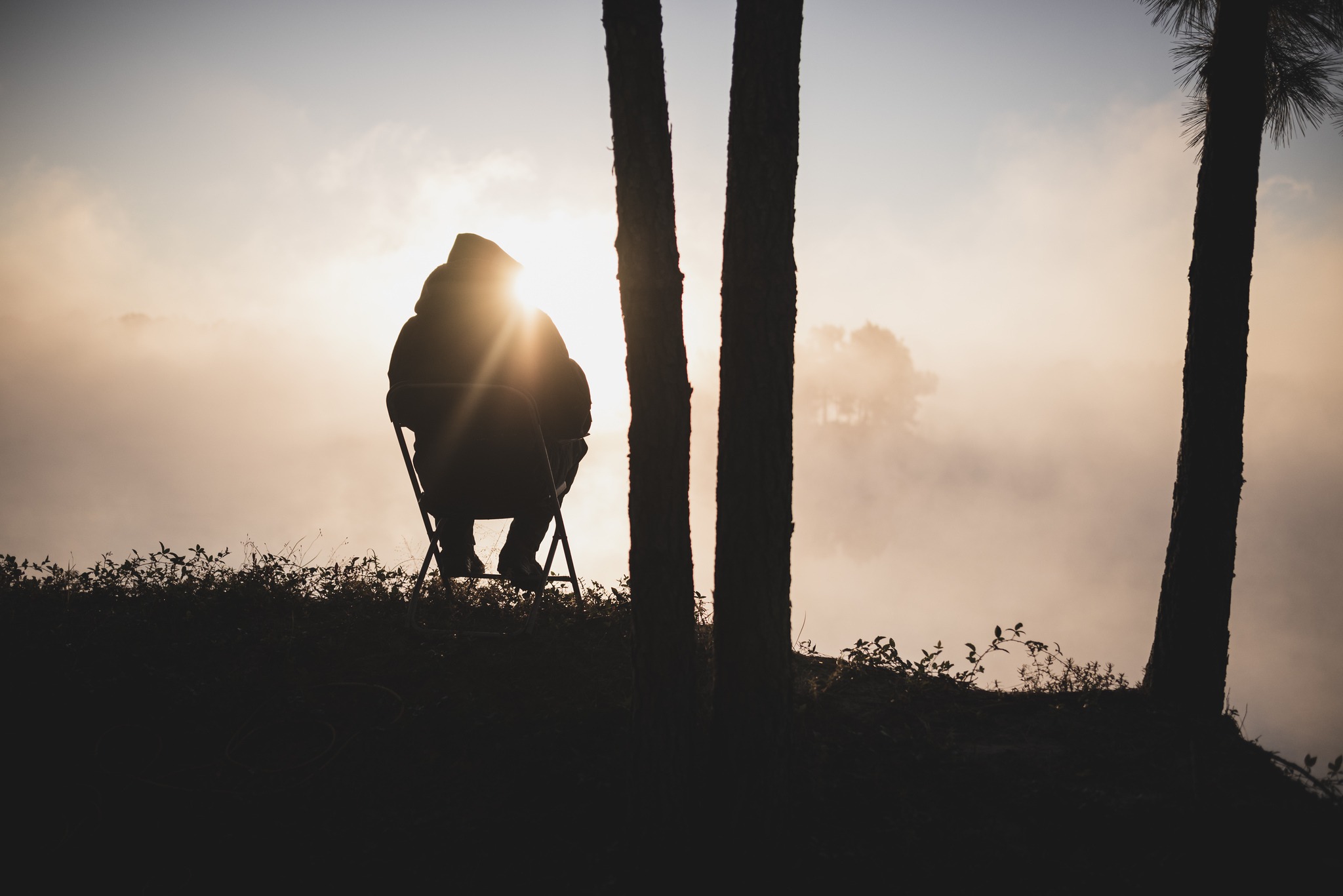 Silhouette of a man seated alone in a folding chair by a misty lake at dawn, sunburst behind him.
