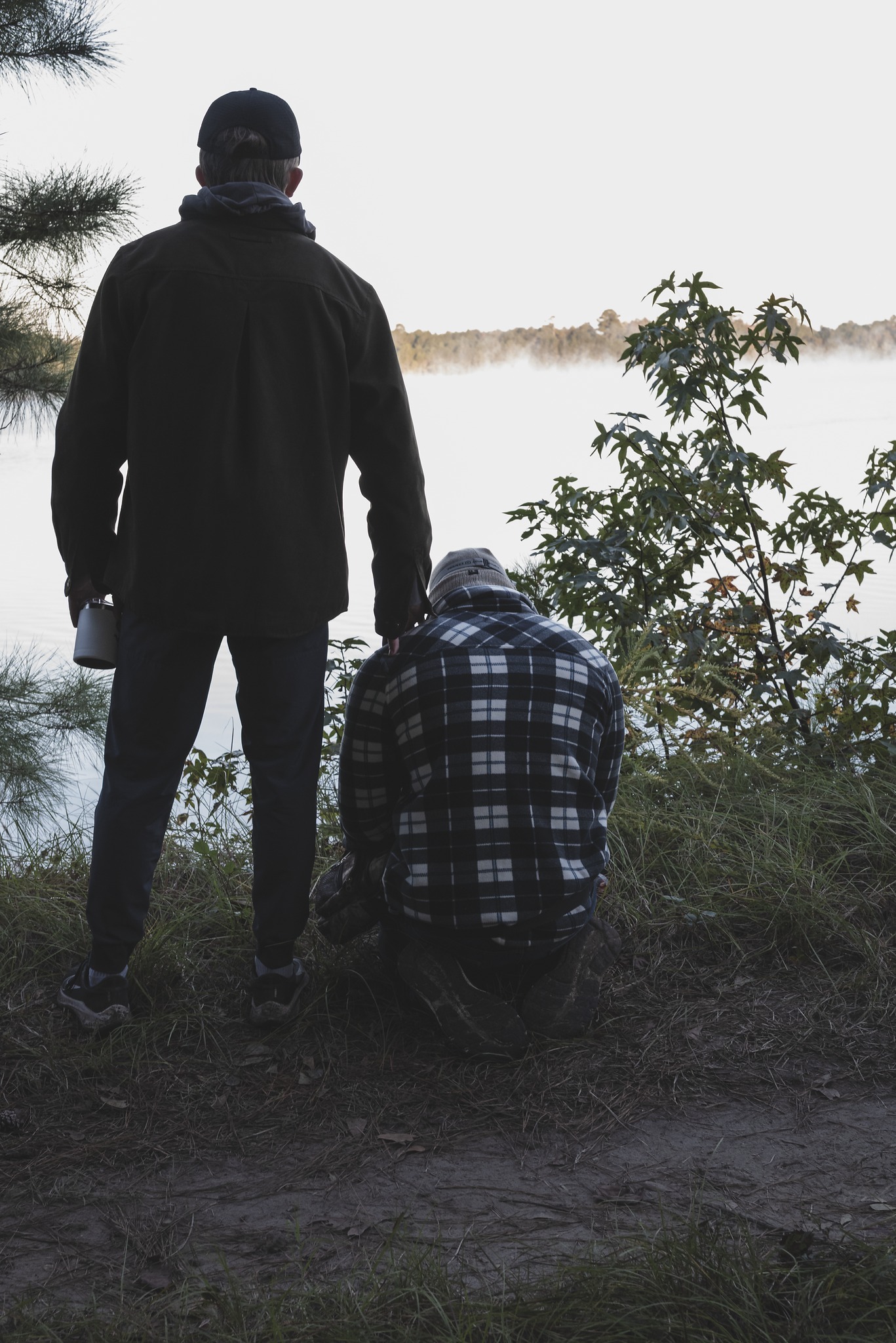 Two brothers at the edge of a misty lake at dawn: one standing with a hand on the other's shoulder.